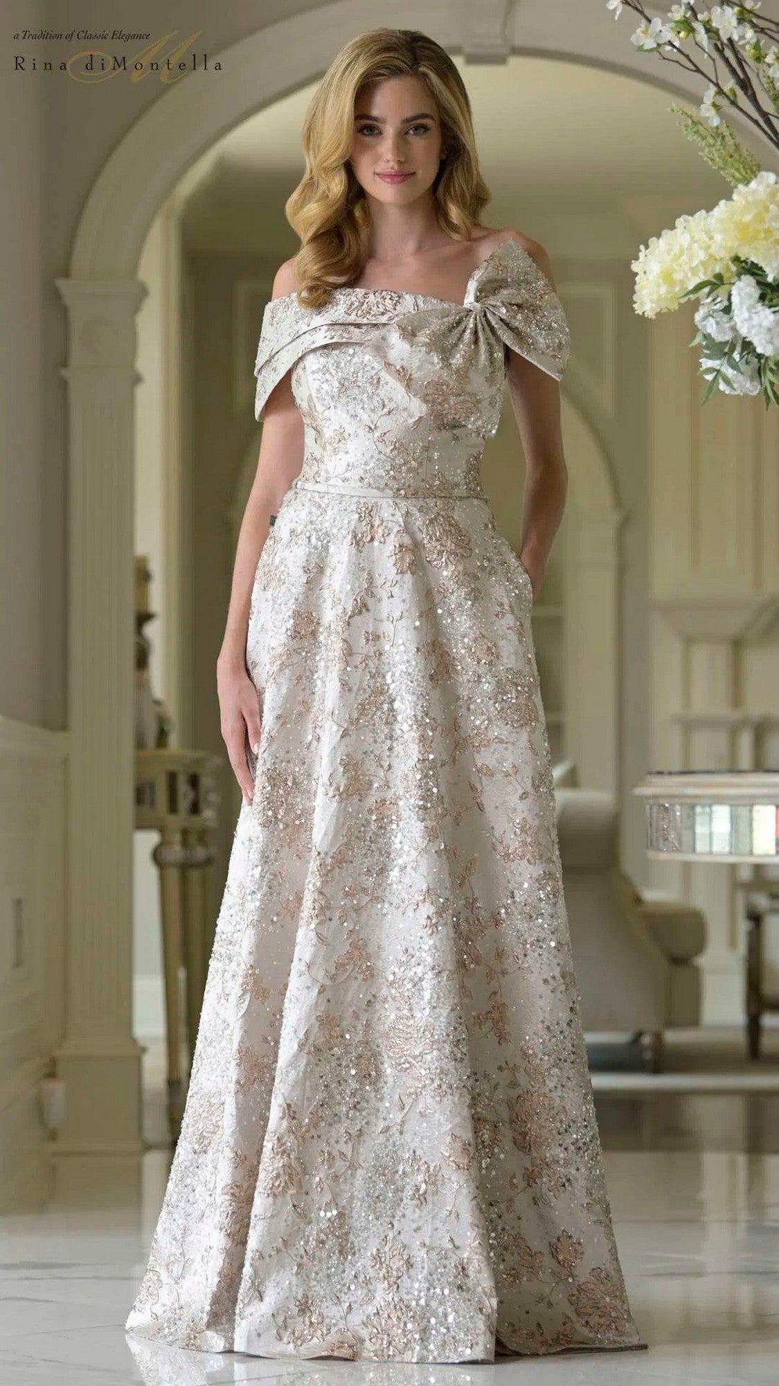 A woman in a gold Rina Di Montella RD3425 long brocade formal dress with an off-shoulder neckline stands in an elegant, light-filled room with classic decor.
